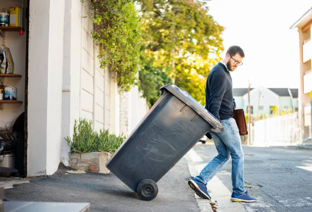 A person wheeling a household bin down a driveway toward the curb on a tree-lined street.
